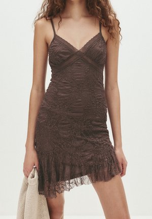 Femme portant une robe en dentelle marron foncé ajustée avec de fines bretelles et un ourlet volanté asymétrique, tenant un vêtement en maille beige.