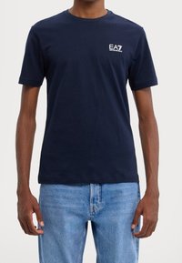 Mörkblå bomullströja med rund hals och korta ärmar. Har en vit EA7-logotyp på vänster bröst. Avslappnad passform, slät textur.