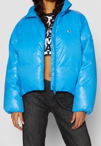 Cropped blue puffer jacket with a shiny texture, front zipper closure, and a small logo on the chest. Long, voluminous sleeves.