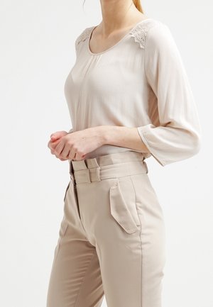 Beige långärmad blus med spetsdetalj på axlarna, kombinerad med högmidjade beige byxor med sidofickor och en mjuk yta.