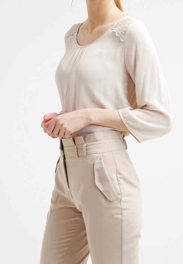Beige langærmet bluse med blonde detaljer på skuldrene, parret med højtaljede beige bukser med sidelommer og en glat tekstur.