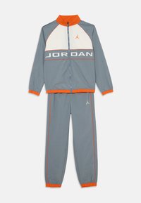 Tuta grigia con accenti arancioni. La giacca presenta un pannello bianco e la scritta "JORDAN". I pantaloni hanno polsini elasticizzati e dettagli con logo. Materiale leggero.