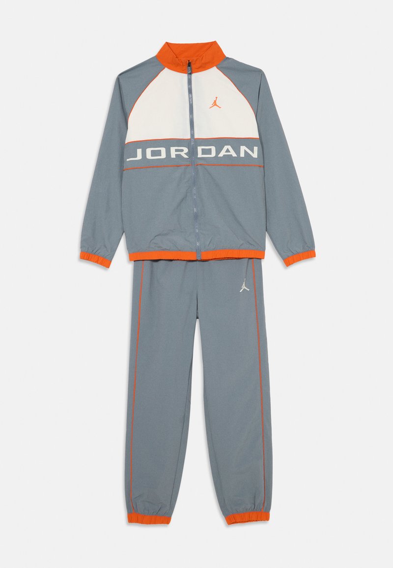 Tuta grigia con accenti arancioni. La giacca presenta un pannello bianco e la scritta "JORDAN". I pantaloni hanno polsini elasticizzati e dettagli con logo. Materiale leggero.