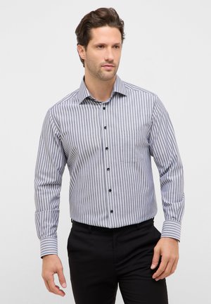 Homme portant une chemise à manches longues rayée verticalement gris et blanc avec des boutons noirs, assortie à un pantalon noir, debout devant un fond uni.