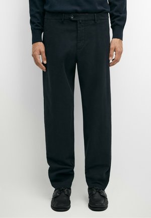 Uomo che indossa pantaloni neri a gamba dritta e scarpe nere con lacci, in piedi davanti a uno sfondo uniforme.