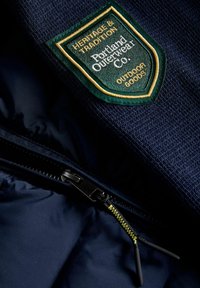 Jūras karafārs ar zaļu un zelta uzšuvi, uz kuras rakstīts "Mantojums un Tradīcija, Portland Outerwear Co." Redzama rāvējslēdzēja detaļa sānos.