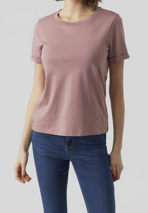 Vrouw in een effen mauve kortärmelig T-shirt en blauwe spijkerbroek, staande tegen een lichtgrijze achtergrond.