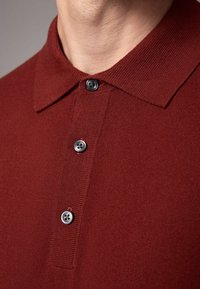 Burgundowa koszulka polo z teksturowanym splotem, z kołnierzykiem w kształcie odwróconego trójkąta i trzema czarnymi guzikami z przodu.