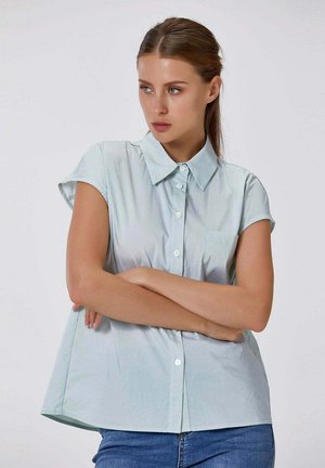 Femme avec chemise à manches courtes bleu clair boutonnée et jean bleu, les bras croisés, regardant sur le côté sur un fond uni.