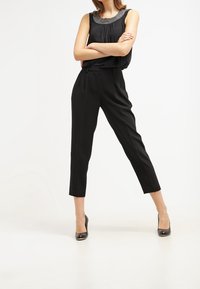 Top nero senza maniche con dettagli in perle lungo il scollo, abbinato a pantaloni neri sartoriali. La modella indossa scarpe con tacco nere.
