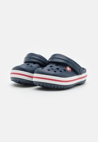 Sabots bleu marine avec des trous de ventilation, semelles rayées blanches et rouges, et brides de talon ajustables sur fond blanc.