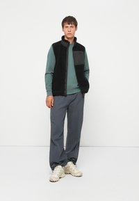 Zwarte fleece vest met een hoge kraag, voorrits en een borstzak, gecombineerd met een turquoise langemouwenshirt en grijze broek, afgewerkt met beige sneakers.