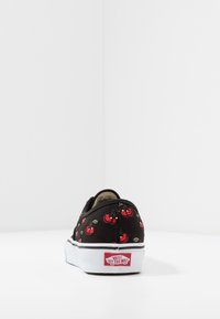 Černé tenisky Vans s červenými cherry vzory, pohled zezadu, opřené o bílou zeď, s viditelným logem značky na patě.