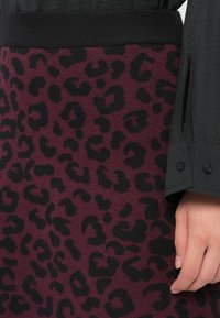 Mano descansando junto a una falda color burdeos con estampado de leopardo negro, combinada con una cinturilla negra acanalada y el puño de una camiseta de manga larga oscura.