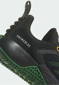 Černá a zelená atletická bota s texturovanou podrážkou, text "PRIMEBLUE" na boku a logo Adidas na jazyku, zobrazená v úhlu v detailním záběru.