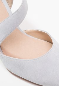 Grå mocka högklackad sko med utskärningsdesign, som har en mjuk textur och ett korsband. Insidan är beige och slät.