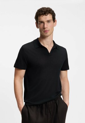 Mann mit kurzen, lockigen Haaren trägt ein schwarzes kurzärmliges Poloshirt und dunkle Hosen, steht mit den Händen in den Taschen vor schlichtem Hintergrund.