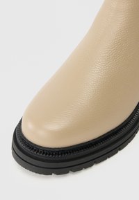 Bottines en cuir beige avec un bout arrondi, surface texturée et semelle crantée noire. Présente une bordure décorative noire le long de la semelle.