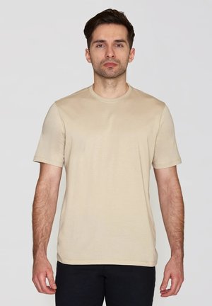 Uomo con capelli corti e scuri che indossa una t-shirt beige semplice a maniche corte e pantaloni neri, davanti a uno sfondo chiaro.