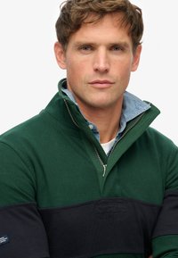 Pullover verde scuro con design a mezza zip, caratterizzato da una fascia a coste blu navy sul petto e un colletto azzurro sovrapposto.
