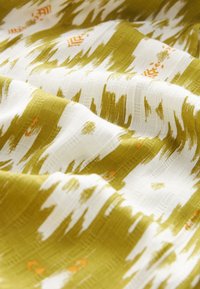 Tissu présentant un motif texturé vert olive et blanc, avec des accents orange. Le tissu semble doux et léger, avec un léger éclat.