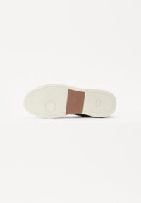 Barna sneaker, texturált bőr felsőrésszel és bézs gumitalppal, amelyen barna logót módosító folt található a sarkán.