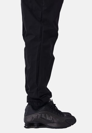 Zapatilla deportiva negra con parte superior texturizada y suela de goma, combinada con pantalones negros ajustados que cuentan con puños elásticos en los tobillos.