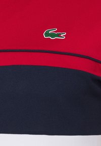 Punainen ja laivastonsininen raidallinen poolopaita, jossa on teksturoitu viimeistely ja pieni vihreä krokotiililogo vasemmassa yläkulmassa.