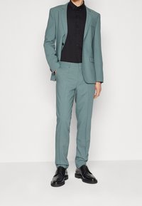 Hellblauer Anzug mit schlanker Passform, kombiniert mit einem schwarzen Hemd mit Knöpfen. Schwarze elegante Schuhe. Das Material wirkt glatt und ohne sichtbare Muster.