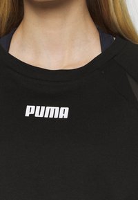 Kvinna som bär en svart Puma-tröja, visar sitt halslinje och övre bröst med synligt blont hår.