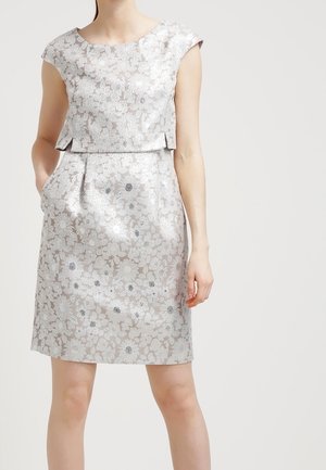 Femme portant une robe courte argentée à motifs floraux avec des poches, debout sur un fond clair uni.