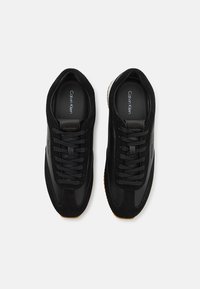 Black sneakers featuring a mix of smooth and suede textures, round laces, and a gum rubber sole. Logo displayed on the insole.