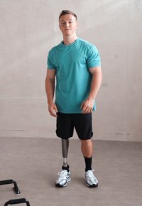 Hombre vestido con una camiseta atlética color teal y pantalones cortos negros, de pie en el suelo de un gimnasio. Tiene una pierna prostética plateada y zapatillas blancas.