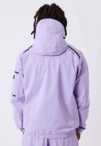 Veste imperméable de couleur violet clair avec une capuche, présentant des accents noirs sur les épaules et les manches, texture lisse et ourlet élastique.