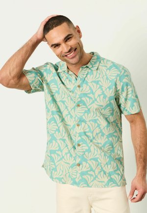 Mann lächelt und berührt sein kurzes Haar, trägt ein hellblaues kurzärmeliges Hemd mit tropischem Muster und beige Hosen.