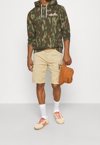 Hoodie kamuflażowy w kolorach zielonym i brązowym, beżowe krótkie spodnie typu cargo, pomarańczowy kapelusz typu bucket, białe skarpetki oraz kremowe sneakersy z czerwonymi akcentami.