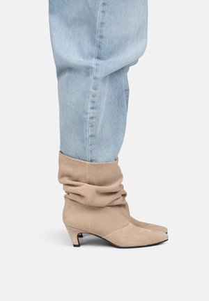 Beige suede slouchy ankle boots with low heels worn with light blue denim jeans against a plain background.