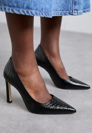 Scarpe con tacchi alti neri con punta affusolata, realizzate in materiale testurizzato che ricorda la pelle di coccodrillo, con un tacco snello e lati aperti.