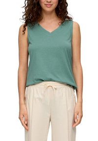 Donna che indossa un top verde senza maniche con scollo a V e pantaloni beige chiaro con cordino, in piedi contro uno sfondo bianco.