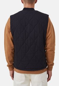 Granatowa pikowana kamizelka z prążkowanym kołnierzem, teksturowanym wzorem i bez rękawów, noszona na brązowej bluzce z długim rękawem.