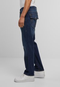 Donkerblauwe denim jeans worden zijwaarts getoond, met een rechte pijp, achterzakken met stikdetails en een comfortabele pasvorm.