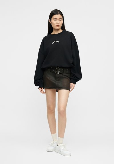 Γυναίκα που φοράει υπερμεγέθη μαύρο φούτερ με λευκό κείμενο, μαύρη δερμάτινη μίνι φούστα με φαρδιά ζώνη, λευκές κάλτσες και λευκά αθλητικά παπούτσια, στέκεται ακίνητη.