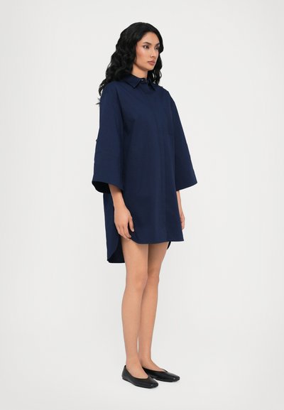 Abito camicia blu navy con colletto, vestibilità ampia, maniche corte e spacco laterale. Il materiale sembra liscio e leggero.