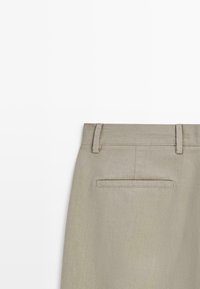 Bakifrån av beige byxor som visar midjeband med bältesöglor och en enkel påsficka på enfärgad bakgrund.