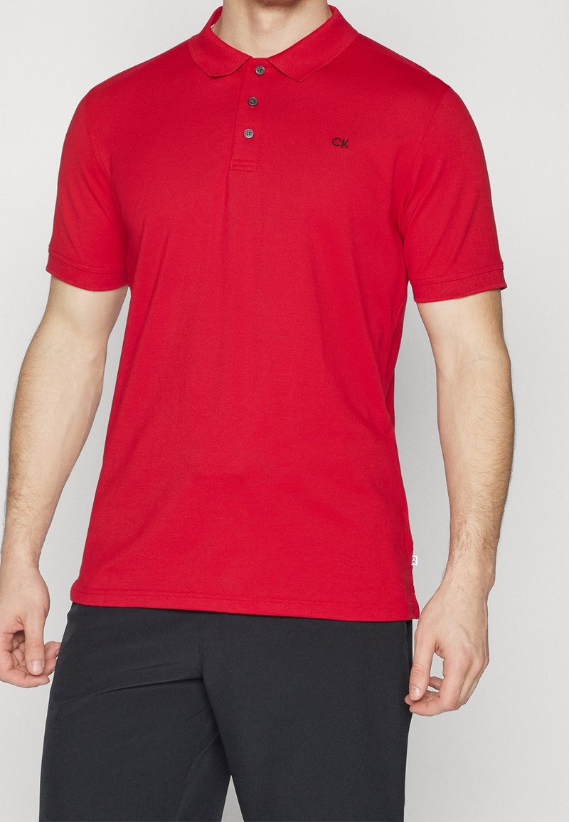 Homme portant un polo rouge à manches courtes avec des boutons noirs et un pantalon noir, debout devant un fond clair uni.