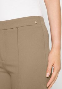 Pantaloni marroni slim-fit con una texture liscia, dotati di una vita piatta, cuciture laterali e un piccolo logo metallico nella parte anteriore.
