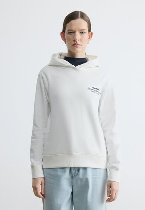 Kobieta w białej bluzie z kapturem GANT z logo na piersi i jasnoniebieskich dżinsach, stojąca na jednolitym jasnym tle.