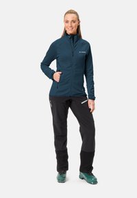 Marine blauwe ritsjack met textuurstof, hoge kraag en ritszakken, gecombineerd met zwarte broek en turquoise skibotten.