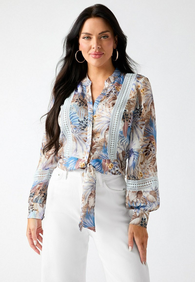 Femme aux longs cheveux foncés portant de grandes boucles d’oreilles en anneau, un chemisier à motif floral avec des détails en dentelle, noué à la taille, et un pantalon blanc.