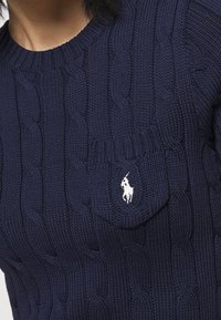 Navy knitted sweater featuring a cable pattern, round neckline, and left chest pocket with a white embroidered logo.